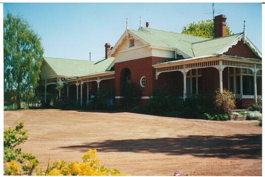 VIctorian style red-brick single story house, green pitched tin roof, deep verandahs, tree and some garden visible.