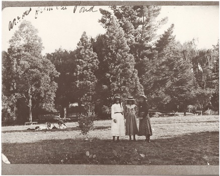 Buninyong Botanic Gardens with three girls standing centre in calf-length dresses, straw hats 1914.  Three more girls lying on grass to left,