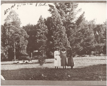 Buninyong Botanic Gardens with three girls standing centre in calf-length dresses, straw hats 1914.  Three more girls lying on grass to left,