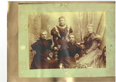 Formal photo of four mature Victorian women, three seated, one standing, all in mutton sleeved dresses, three in black, lady on left in lighter colours.