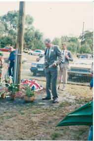 Gentleman in grey suit, holding paper, standing next to pole, low memorial covered with Australian flag, open ground behind with parked cars and trees beyond.
