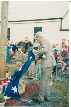 Man in suit removing flag to unveil memorial, people in background including boy scouts, and side of Scotsburn Hall.