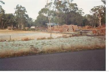 View across road to woodyard, part of field to left, Woodpiles, shed and truck visible, trees behind.