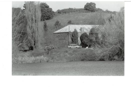 Bluestone, iron-roofed  building, nestled between trees, hill rising behind, reservoir in foreground