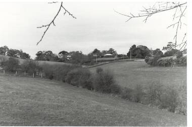 View across fields to house on top of hill, bushes following fence-lines centre, trees along skyline.