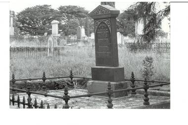 Granite headstone on plinth with triangular cap, wrought iron railing, inscription, other headstones behind, trees.