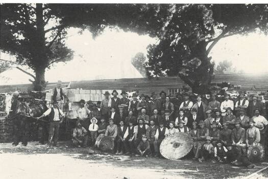 Workmen in three rows, front row seated, next to horse and cart loaded with wooden boxes, three men holding circular saws, low wooden building behind.