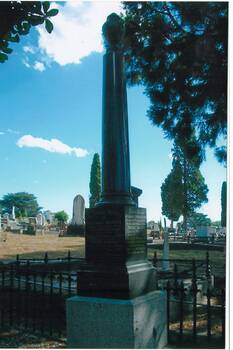 Colour photo, cemetery, monument, polished granite, pillar on engraved plinth, urn cap, cast iron fence, other monuments and trees in background.