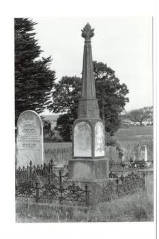 B/W phot cemetery, marble arched engraved headstone on left, centre stone monument, plinth with engraved marble inserts, narrow stone spire with stylized lamp capping.