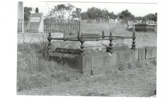 B/W photo, grave with piched lid, cast iron fence on bluestone base, inlaid inscription, "Rennie" headstone visible behind, more headstone in background, overgrown grass.