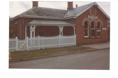 L-fronted brick building, white picket fence, veranda on left with ornate wooden posts, three arched windows on right, polychrome capping and Buninyong Post Office signage.