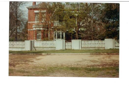 Two story brick house, bay window on left, dou8ble veranda right, obscured by tree, iron lace fence.
