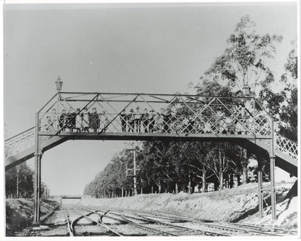 Iron footbridge over railway line, lined with children, anoth bridge crosses the line in the distance, rail tracks in shallow cutting, trees on right.