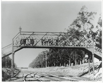 Iron footbridge over railway line, lined with children, anoth bridge crosses the line in the distance, rail tracks in shallow cutting, trees on right.