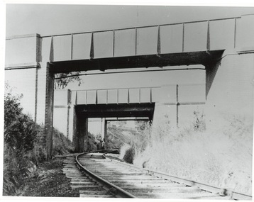 Single rail line curving right through a cutting under three brick and steel plate bridges