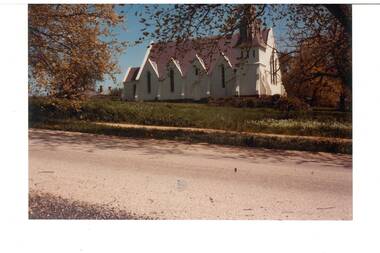 White stucco church, multiple gables and steeple, across unmade road, through trees.