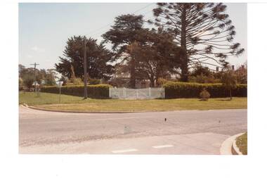 View across intersection. white picket gate in hedge, large trees beyond in garden obscure the house.