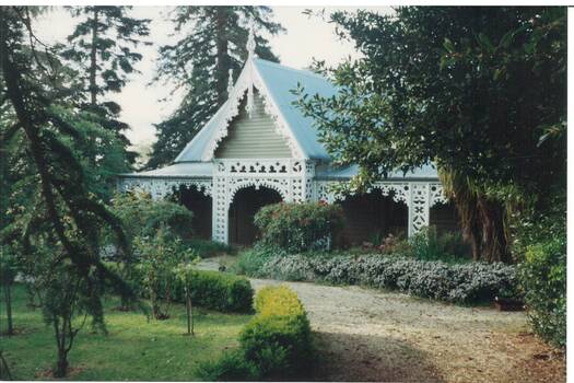 Weatherboard house, high gable, ornate carved wood barge-boards and veranda lace-work. Lush garden