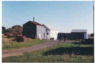 Iron and wood farm buildings, tin roofs, two men walking along path towards buildings