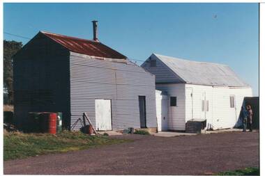 Two farm buildings, one grey tin, other white board, tin roofs, gravel path.