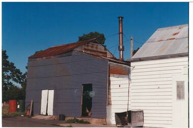 Side view, grey tin farm building, open arch and pair of rough white doors, peaked tin roof, part of adjoining white weatherboard building, some barrels.