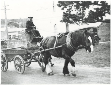 Old manual horse-drawn fire engine in poor condition with man seated in uniform driving horse uphill, shops in background