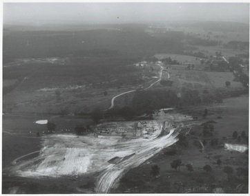 Aerial view, fields and forest with roadway curving across centre, large earthworks, clearing and grading in foreground.