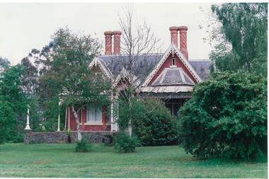 Brick house on raised bluestone foundations, triple gables with carved barge boards, verandah with iron lace, two pairs of chimneys, brick in candy-cane pattern.