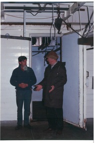 Two men in cold room, white glossy insulated walls and door, pipes, rails and hooks for moving carcasses above.