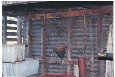 Inside corrugated iron shed, wooden frame, tubs, chains, funnel, hand-pump, piping.