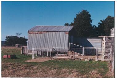 Large raised  tin shed, ramp down into holding pen, pipe and wire fencing, wood fences behind, and part of wooden building to one side.