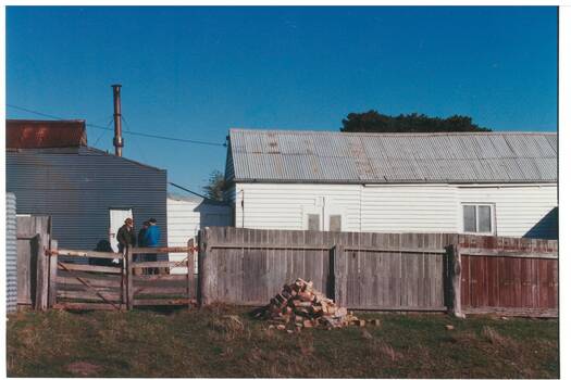 Side view two sheds, one tin, one wood, behind wooden fence. Pile of bricks centre.