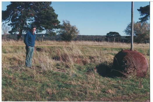 Man in blue jumper standing in field near old rusted boiler, trees and bush in background