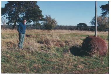 Man in blue jumper standing in field near old rusted boiler, trees and bush in background