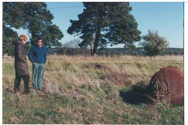 Two men in field, one in coat and deerstalker hat, other in blue jumper, rusty boiler nearby, trees and bushland beyond.