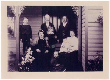B/w family photo on veranda, small girl sitting on ground, two ladies, one holding a baby, two boys and two men behind.