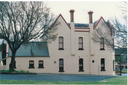 Two story brick building, tall windows, painted cream, two high gables, two chimneys with ornate brickwork. Single story extension on left.