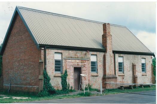 Plain brick building, central chimney, four high windows, simple door between two left-side windows. Part of tennis court, net and umpires stool in foreground.
