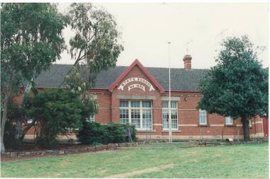 View centre of single story brick building, high gable, "State School" signage, large square windows, shrubs and trees foreground at sides.