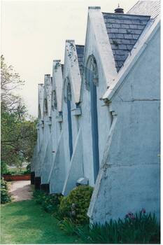 Wall of whitewashed render, showing six buttresses and five gables with tall narrow arched windows, garden, lawn and path on side.