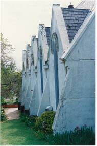 Wall of whitewashed render, showing six buttresses and five gables with tall narrow arched windows, garden, lawn and path on side.