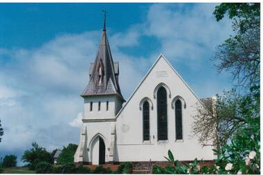 Church, whiewashed render, slate-clad steeple on left over gothic entrance, on right high peaked wall, three tall narrow stained glass windows.