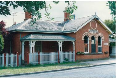 L-fronted single story brick building, veranda with wooden posts and picket fence, three arched windows and Post Office signage, two square chimneys.