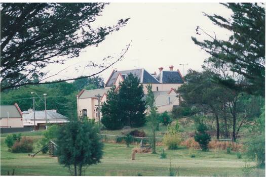 View across parkland to collection of buildings, old with slate roof surrounded by new with iron roof, ground sloping up to right.