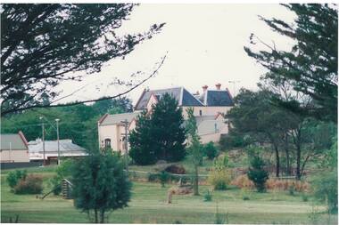 View across parkland to collection of buildings, old with slate roof surrounded by new with iron roof, ground sloping up to right.
