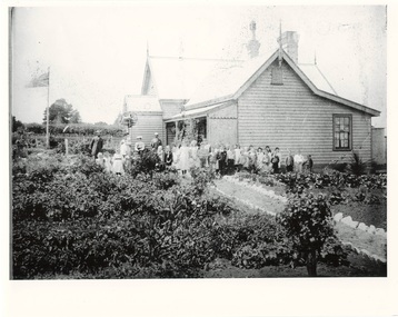 Wooden schoolhouse side view, high gables, verandah, 30 or so children lined up with teachers in well tended garden.