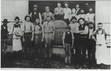 17 men and boys in shirtsleeves standing in doorway and in front of shed, around bale of wool labelled "Larundel'.