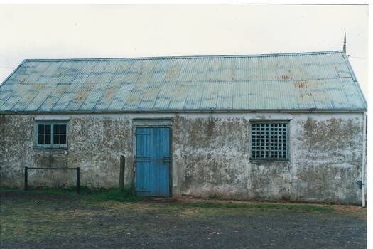 Single story stucco building, traces of whitewash, tin roof, traces of blue paint, blue door centre, windows either side, one grilled.