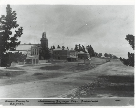 Looking across dirt road, various buildings, town hall centre with tower, small toll-booth shed, horse and buggy middle of intersection.