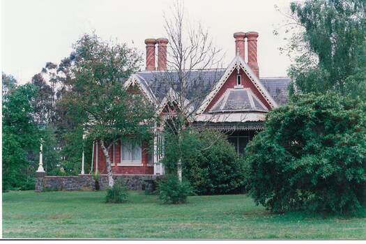 Front view tall brick house in high stone foundation, high orate gables, partially blocked by trees.